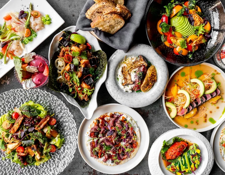 A vibrant assortment of gourmet vegetarian dishes beautifully arranged on various plates and bowls. The meal includes fresh salads, roasted vegetables, and plant-based mains, accompanied by bread. The presentation is elegant and colourful, showcasing a variety of textures and flavours.