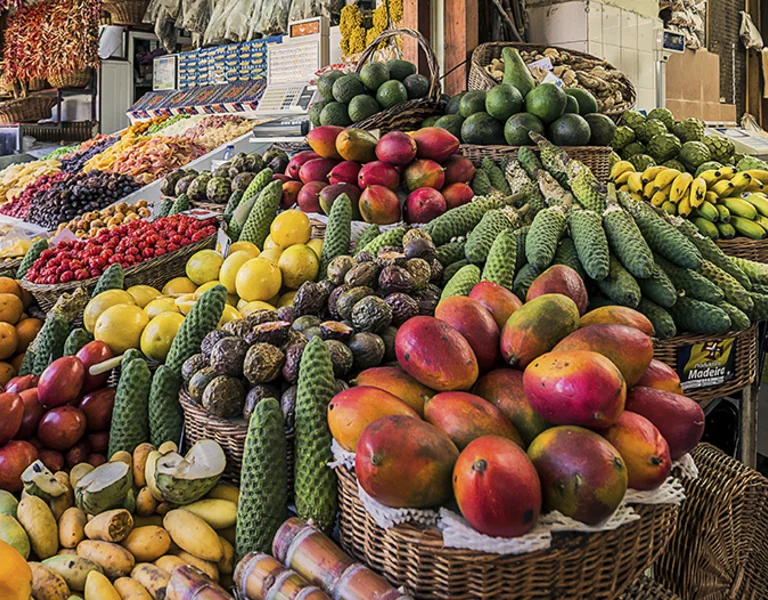 A bustling fruit and vegetable market stall in Madeira, featuring an abundance of colourful produce, including mangoes, bananas, avocados, and exotic fruits. The display is rich and inviting, showcasing local agricultural diversity.