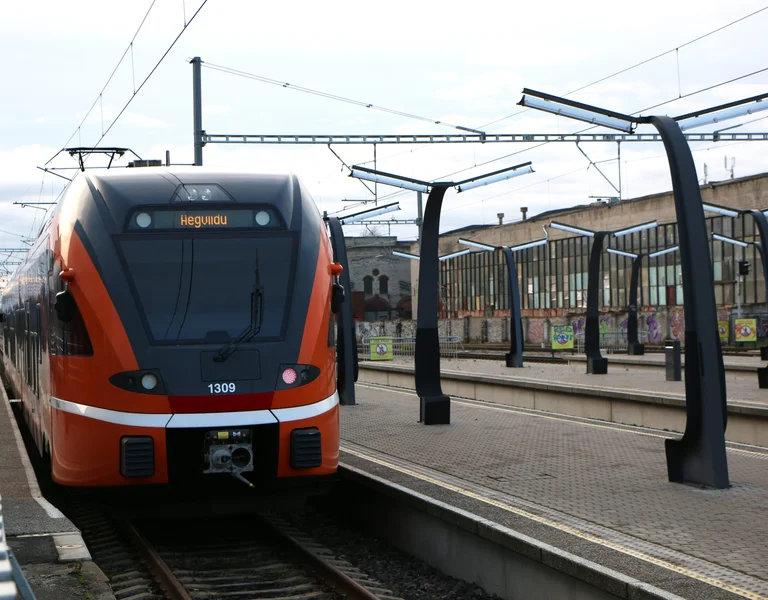 A red train arriving at a station platform, with overhead wires and clear skies.
