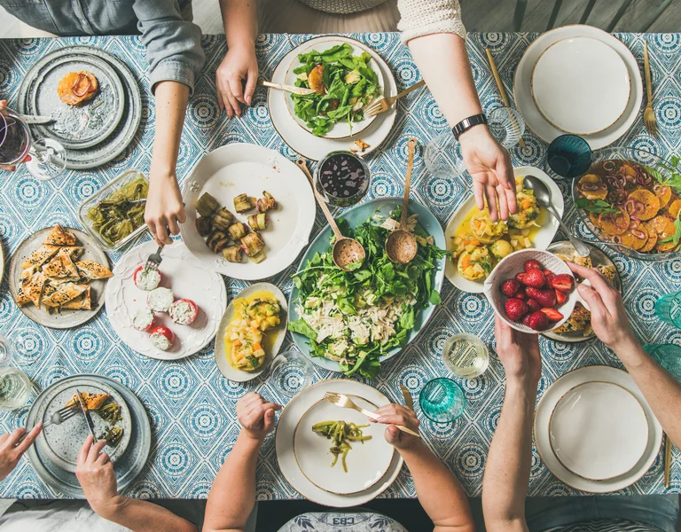A view of a shared meal, featuring various vegetarian dishes on a patterned blue tablecloth. People are seen reaching for food, sharing plates, and pouring drinks, creating a lively, communal dining experience.
