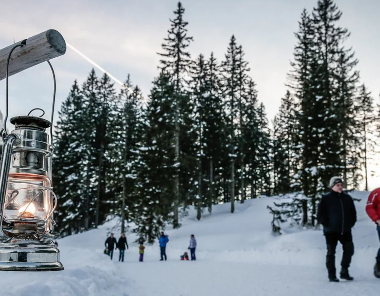 A lit lantern in the snow with people walking in a snowy forest background.