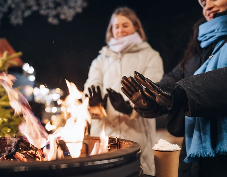 Two people warming hands over an outdoor fire pit in winter, with a Christmas tree in the background.
