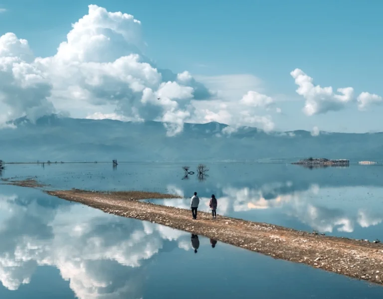 Two people walking on a pathway dividing a lake with mountains and clouds on the background.