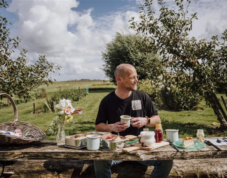 Person at a rustic outdoor table with a picnic setup, holding a cup, greenery in the background.
