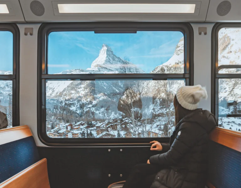 A passenger sits comfortably inside a train, gazing out through a large panoramic window at a breathtaking winter landscape. The iconic Matterhorn mountain dominates the view, framed by a clear blue sky and snow-covered rooftops nestled in a picturesque a