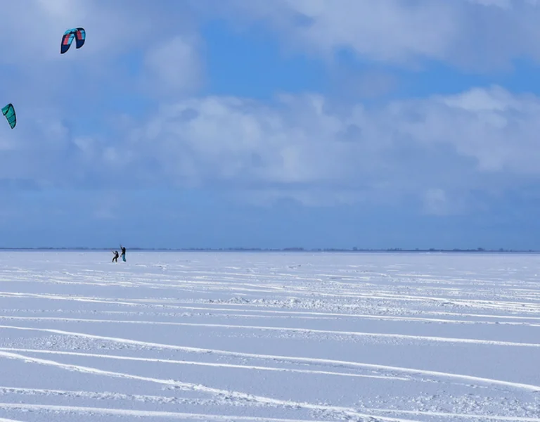 Four snowkiters on a frozen landscape with colorful kites against a cloudy sky.