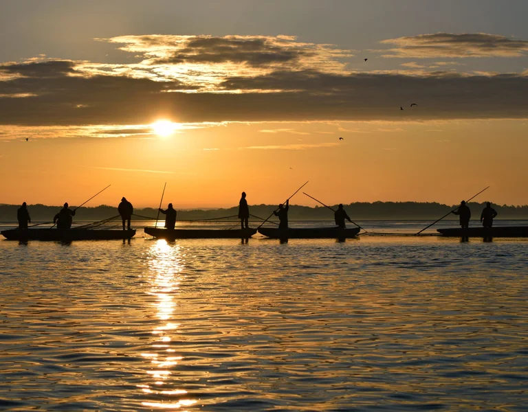Fishermen on boats silhouetted against a golden sunrise over calm waters.