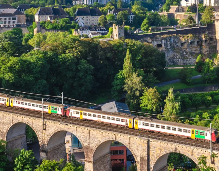 A train crossing an arched viaduct among lush greenery with Luxembourg city buildings in the background.