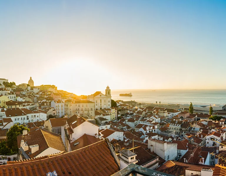 Panoramic view of Lisbon at sunset with orange rooftops and a church.