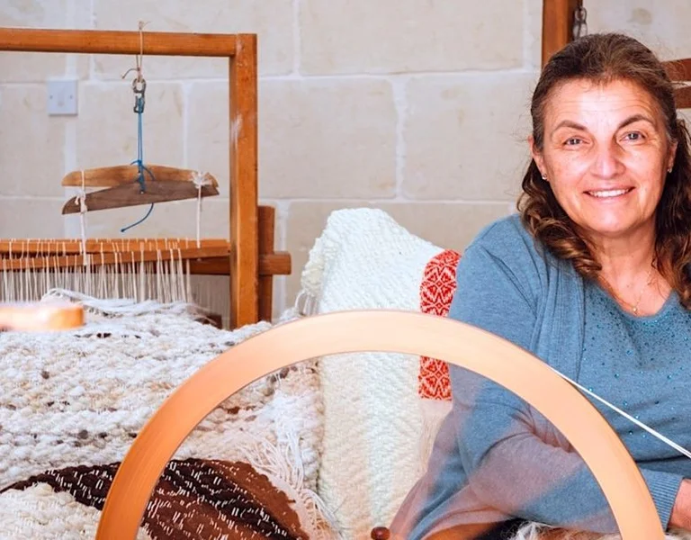 A woman sits at a spinning wheel, surrounded by woven textiles and wooden weaving equipment in a craft studio.