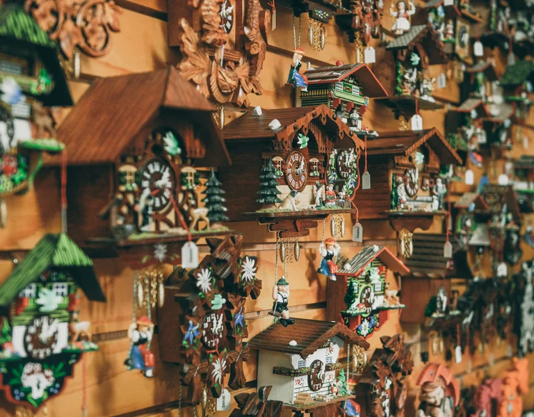 A display of traditional Black Forest cuckoo clocks hanging on a wooden wall. The handcrafted clocks feature intricate carvings, chalet-style designs, and figurines, with small tags attached to each. The warm lighting highlights the wood tones and details of the clocks.