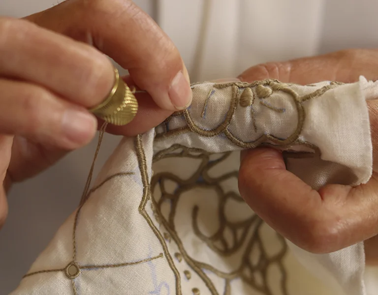 A close-up of hands sewing intricate embroidery on fabric, showcasing detailed craftsmanship and a gold thimble on the thumb.