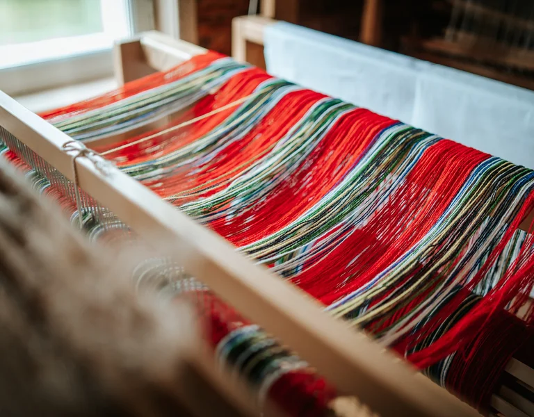 A close-up view of a weaving loom with colorful threads, predominantly red, arranged in vibrant patterns. Natural light illuminates the scene.