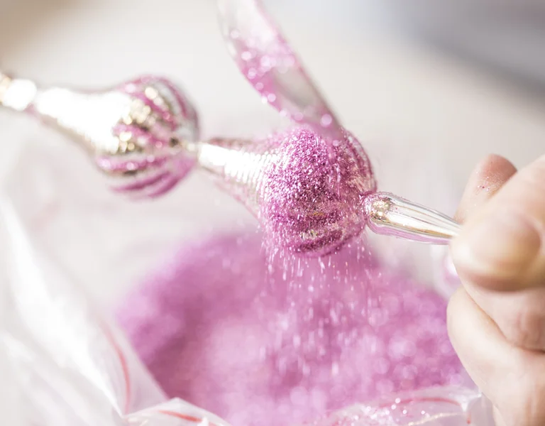 A close-up of a hand sprinkling vibrant pink glitter from a decorative tool over a bag of pink glitter.