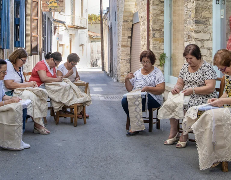 A group of women sit on wooden chairs in a cobblestone alley, engaged in crafting delicate lacework. Vibrant shopfronts line the street.
