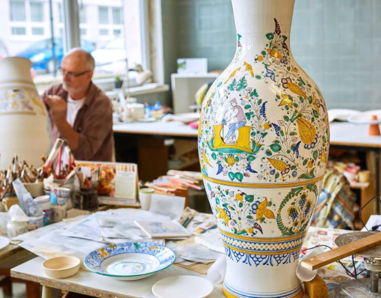 A brightly painted ceramic vase adorned with intricate floral designs sits on a cluttered art studio table, surrounded by various tools and materials.