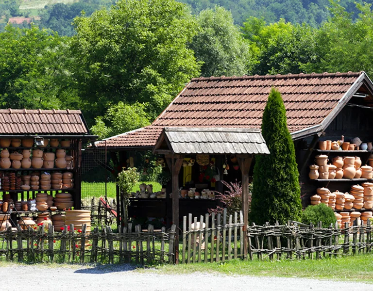 Rustic pottery market featuring wooden stalls filled with clay pots, surrounded by lush greenery and a wooden fence.