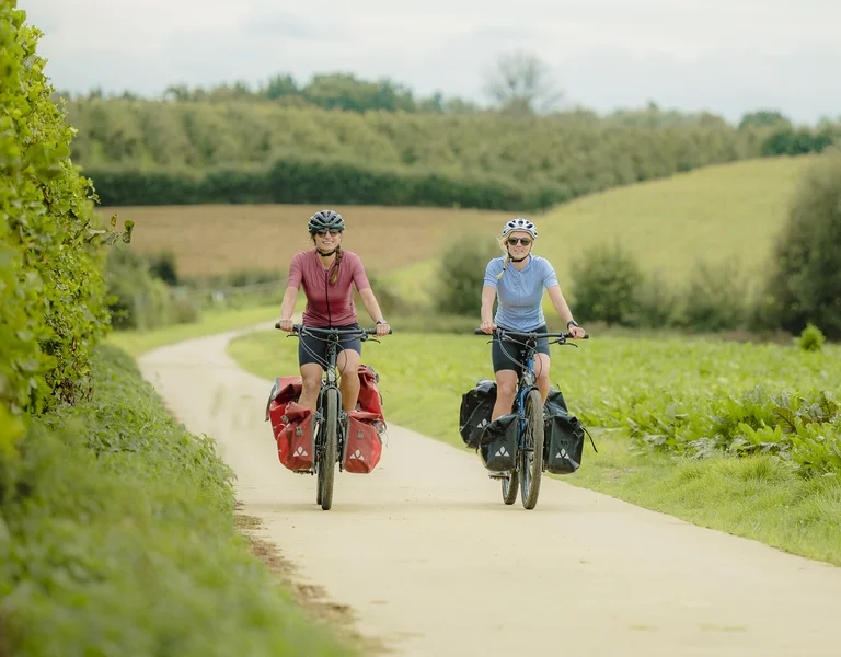 Two cyclists ride along a scenic path, surrounded by lush greenery and farmland, each with loaded panniers on their bikes.