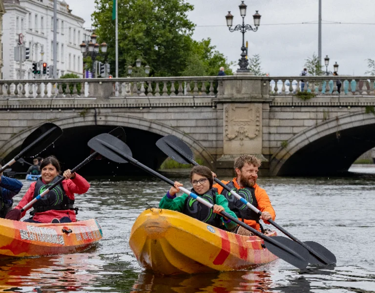 Four individuals kayak on Dublin river surrounded by historic buildings and a stone bridge under a cloudy sky.