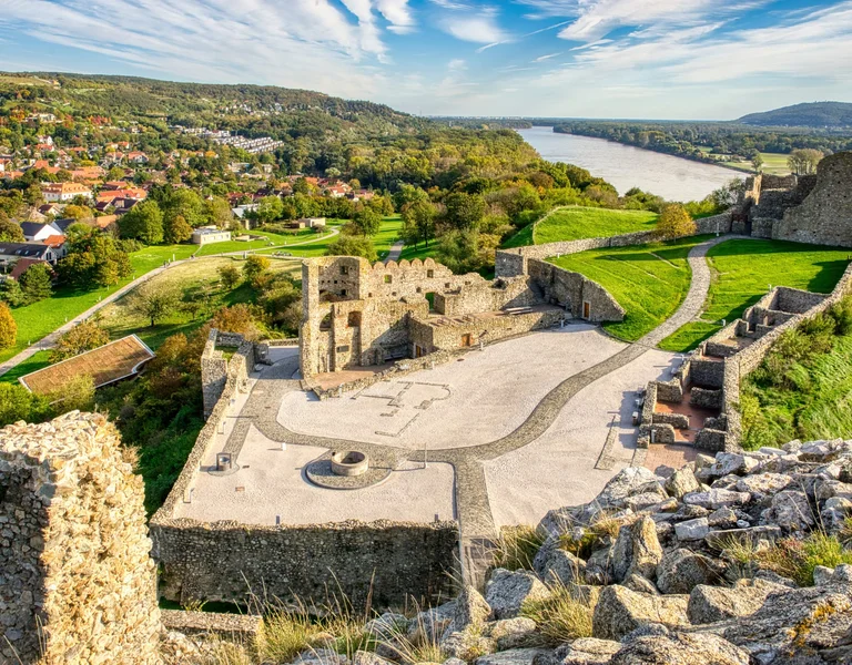 A medieval stone castle on a hilltop overlooking the Danube River, with surrounding green landscapes and village views.