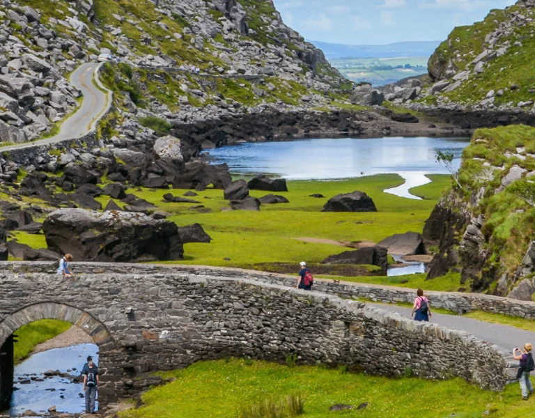 A stone bridge spans a tranquil stream, surrounded by lush greenery and rocky hills, with hikers strolling along scenic roads.