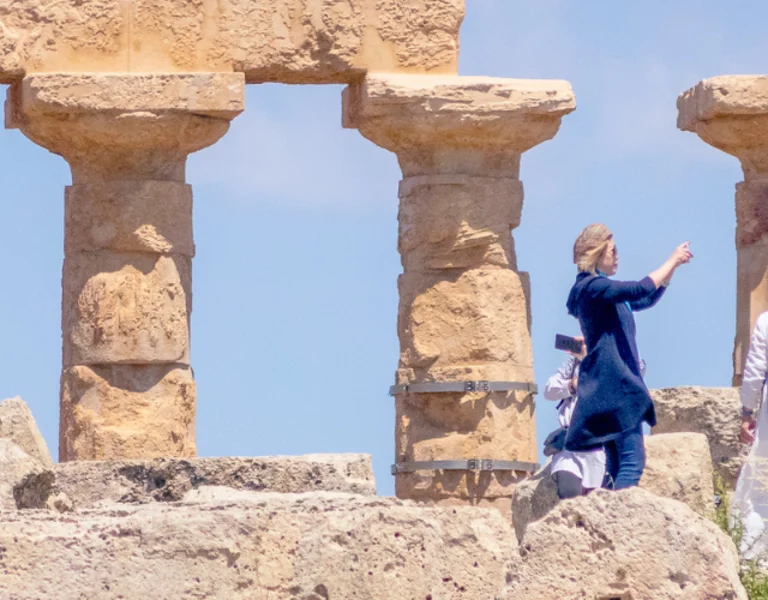 Visitors explore ancient ruins with stone columns amidst lush greenery and a clear blue sky.