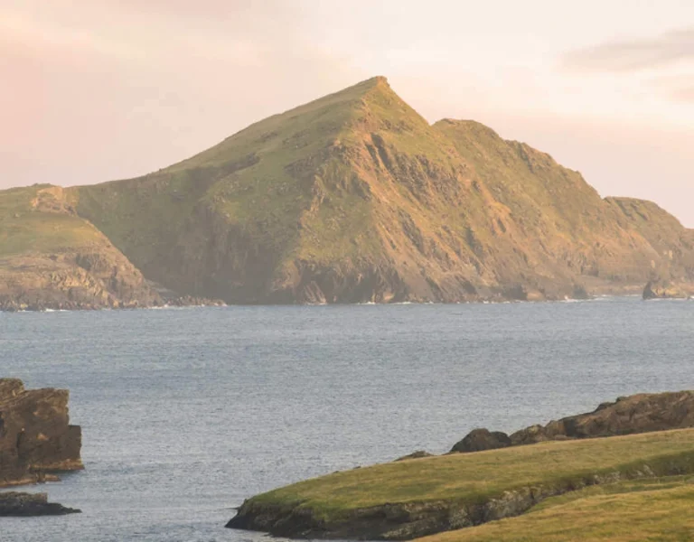A lush green hill rises sharply from the sea, bathed in soft, pastel sunset light, surrounded by rocky shores and calm waters.
