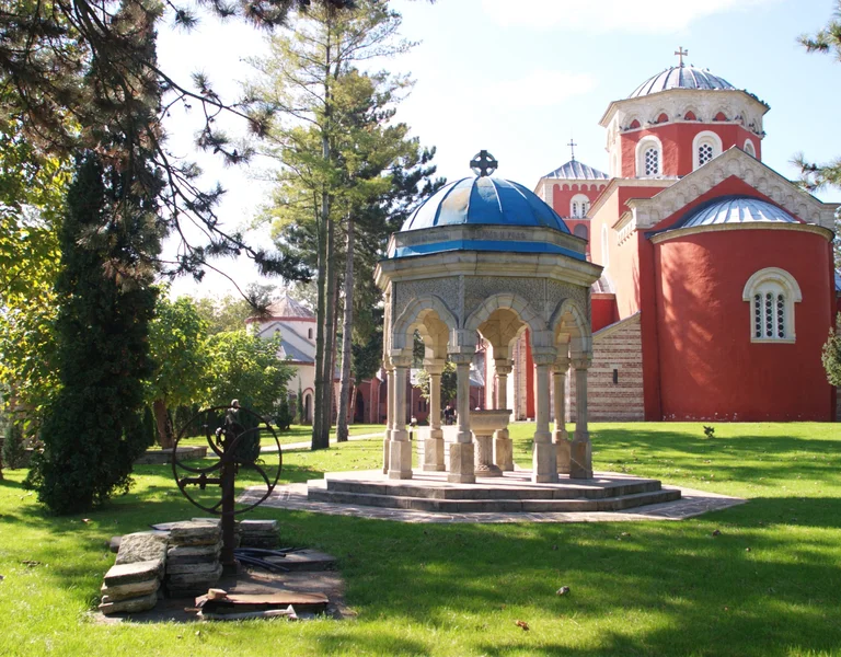 An Orthodox monastery with vibrant red walls and domed roofs, surrounded by green gardens and a stone gazebo.