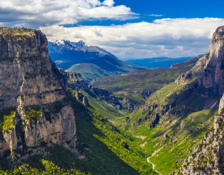 Breathtaking view of a lush valley surrounded by steep cliffs and distant snow-capped mountains under a partly cloudy sky.