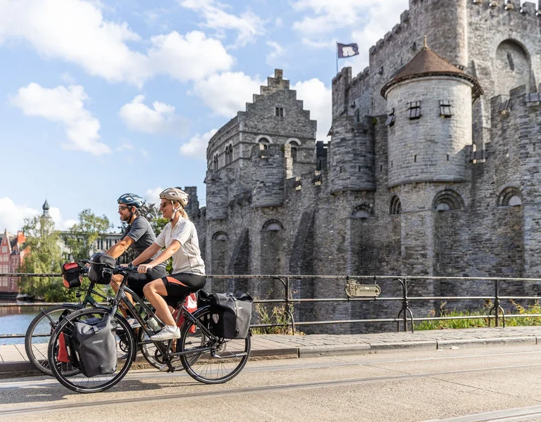 Two cyclists ride along a riverside pathway with a medieval stone castle in the background under a bright blue sky.