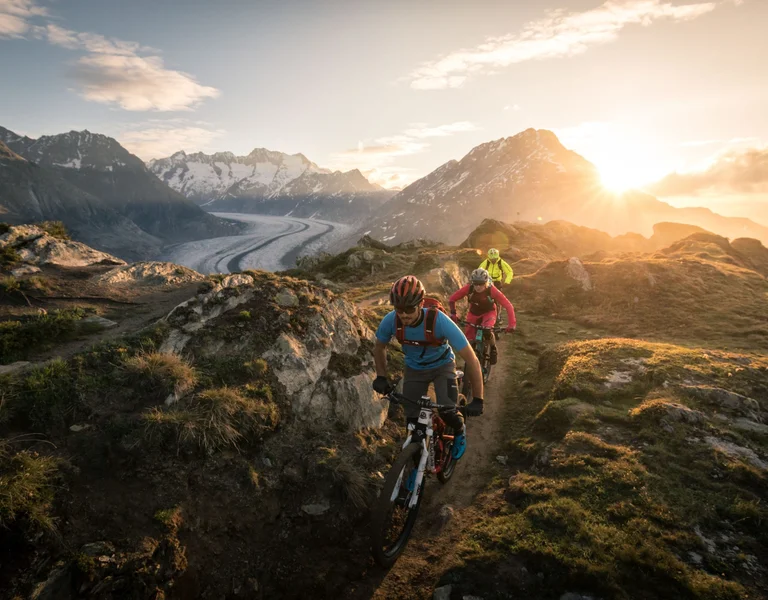 Three mountain bikers ride along a rugged alpine trail at sunrise, with snow-capped peaks and the sweeping Aletsch Glacier in the background.