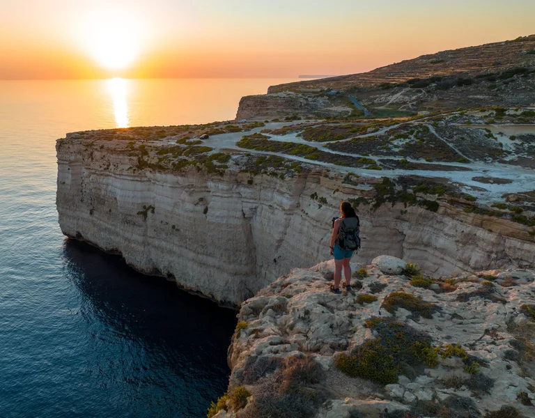 A hiker stands on the edge of limestone cliffs at sunset, overlooking the calm Mediterranean Sea in a rugged coastal landscape.