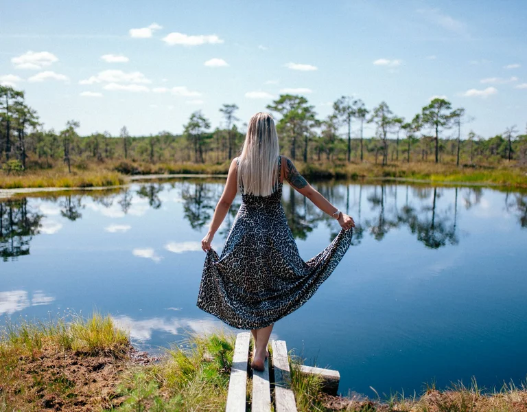 A woman in a leopard-print dress stands on a wooden path beside a calm pond, enjoying a sunny day surrounded by nature.