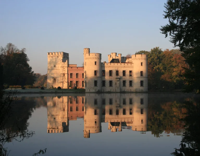 A scenic castle with turrets reflected in a calm lake, surrounded by autumnal trees under a clear blue sky.