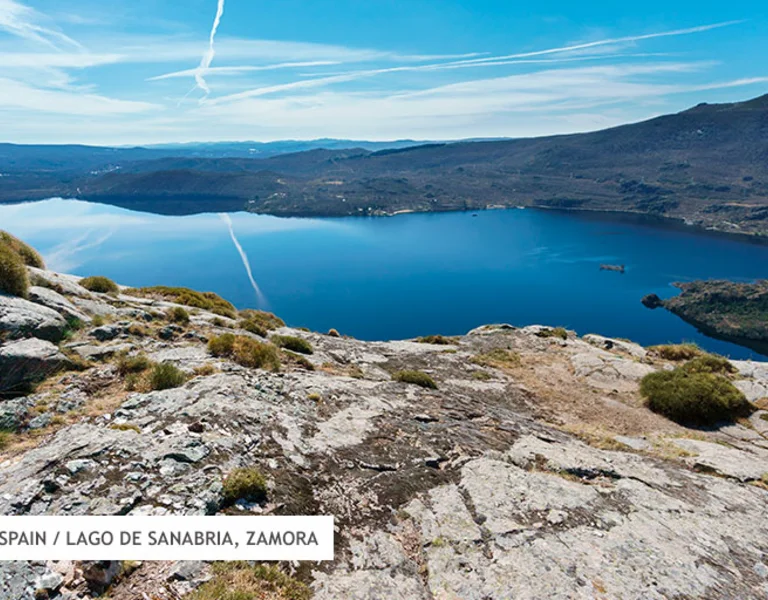 Panoramic view of Lago de Sanabria in Zamora, surrounded by rocky terrain and lush greenery under a clear blue sky.