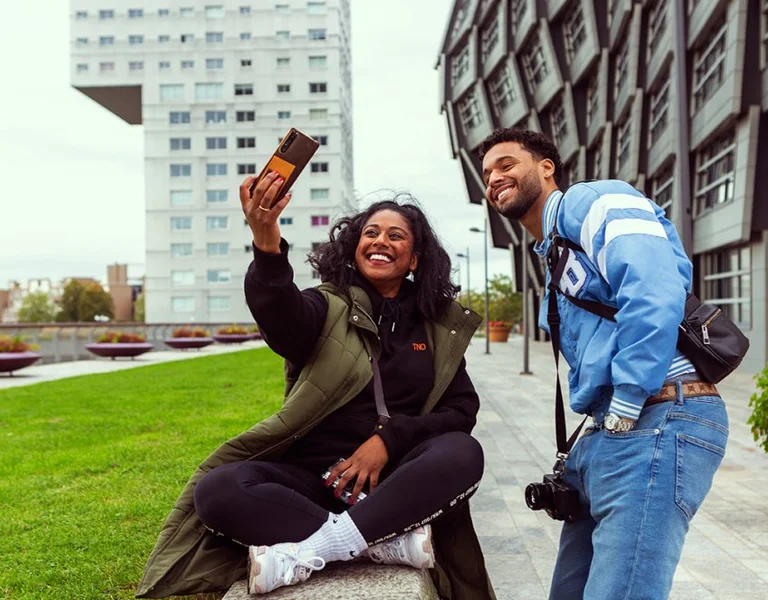 A person sits on the grass posing for a selfie, while another person leans in, smiling, amidst modern architecture and greenery.