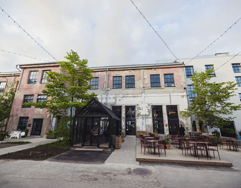 An urban outdoor cafe with rustic charm, featuring an old building, terrace seating, and lush green trees under a cloudy sky.