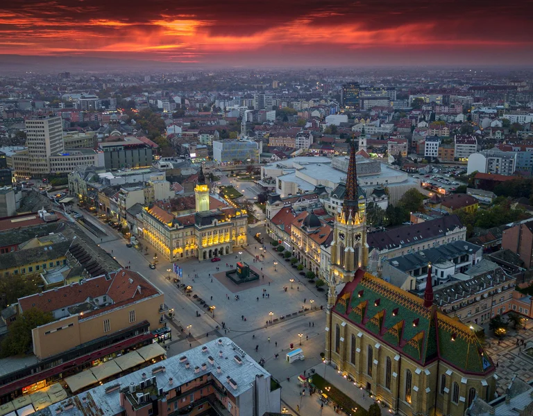 Aerial view of Novi Sad at sunset, featuring the central square, historic buildings, and the Name of Mary Church.