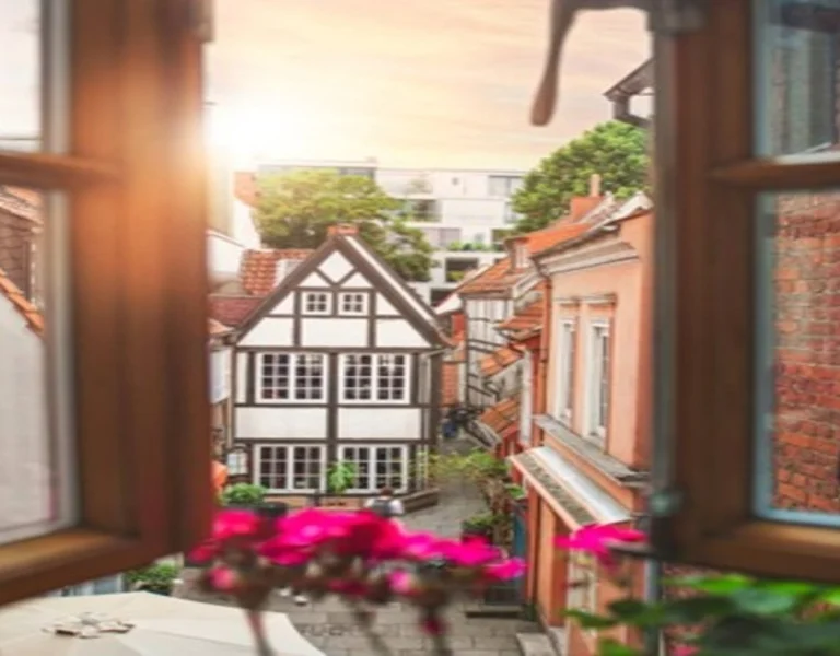 View from a window showcasing a charming street with a half-timbered house and vibrant flowers as the sun sets.