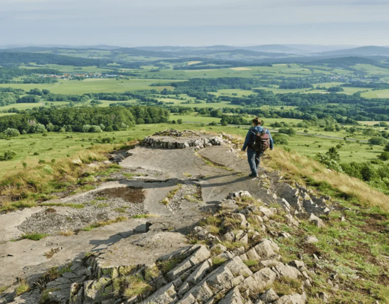 A hiker walks along a rocky ridge, overlooking lush green valleys and distant mountains, under a clear blue sky.