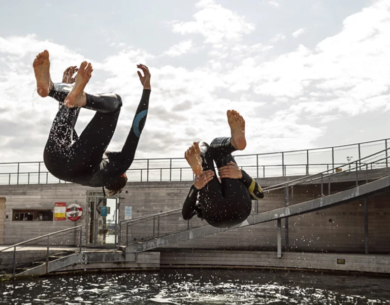 Two surfers in wetsuits dive into a circular swimming area, creating splashes beneath a bright sky and wooden deck.