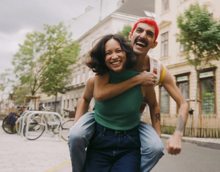 A person gives a piggyback ride along a charming street lined with trees and historic buildings, capturing a joyful moment.