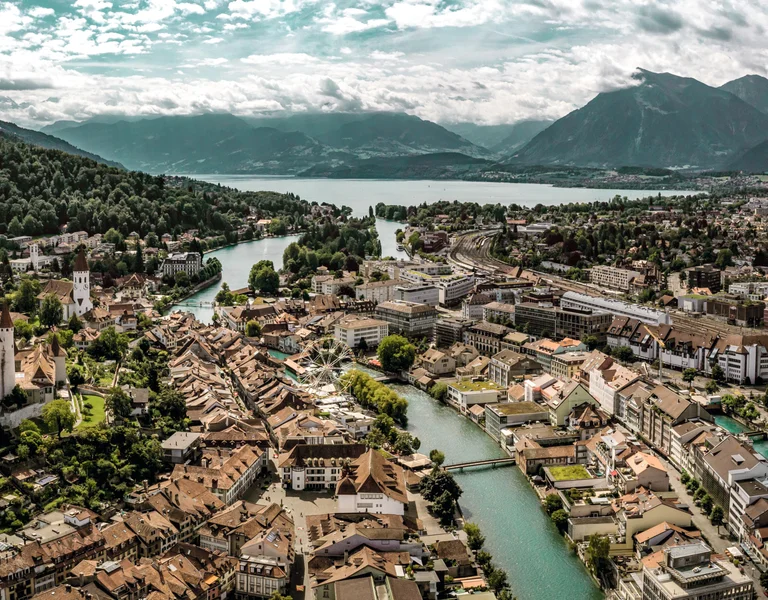 Aerial view of a picturesque town with a historic castle, lush green hills, a winding river, and distant mountains under a cloudy sky.