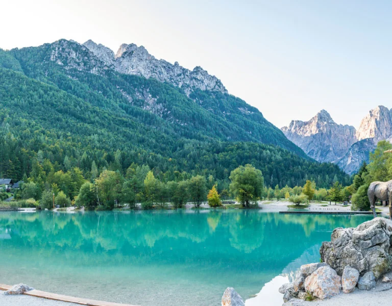 Panoramic view of Jasna Lake in Slovenia, featuring crystal-clear water, surrounding forest, and the iconic ibex statue with the Julian Alps in the background.