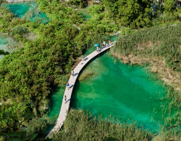 Visitors stroll along a wooden boardwalk, surrounded by lush greenery and vibrant turquoise waters, Plitvice Lakes in Croatia