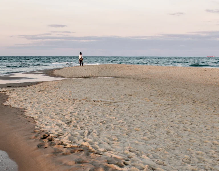 View of Grenen beach in Skagen, Denmark, where the North Sea and Baltic Sea meet, with a person walking along the sandy shoreline at sunset.