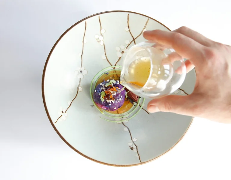 A hand pouring sauce over an artistic plate featuring a vibrant purple dumpling, garnished with colorful edible flowers and seasoning, served in a delicate floral-patterned bowl.