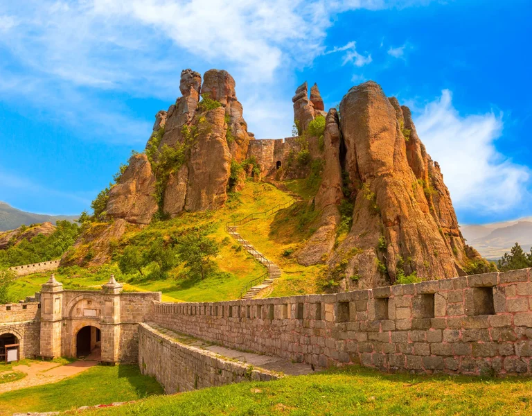 Belogradchik Fortress in Bulgaria, showcasing ancient stone walls and towers atop striking red sandstone formations under a blue sky dotted with some clouds.