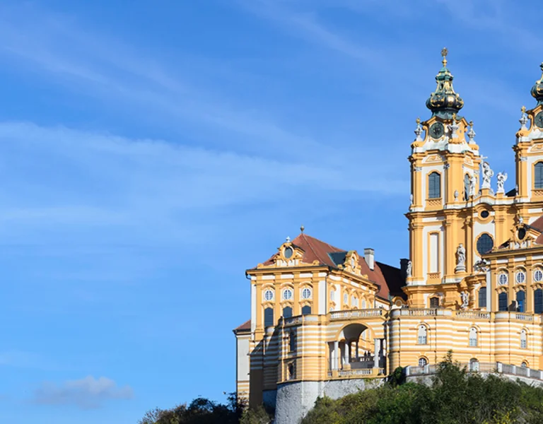 A panoramic view of a majestic yellow and white Baroque monastery, featuring two elaborate towers with green cupolas, perched on a hill against a clear blue sky.