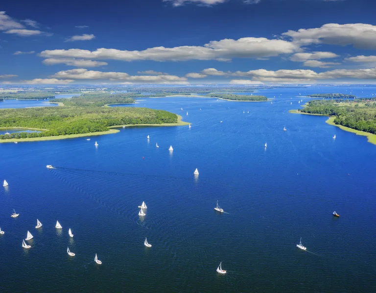Aerial view of the Mazury Lakes in Poland, showing numerous white sailboats on the deep blue water surrounded by green forested islands and banks.